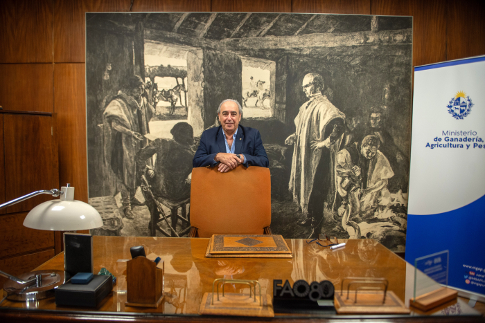 Ministro Fratti, de pie, con los codos sobre le respaldo de una silla. Delante, su escritorio. Detrás, un cuadro con la figura de José Gervasio Artigas