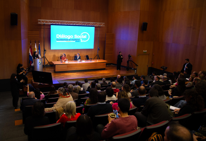 Sala del evento vista desde atrás con público y expositores