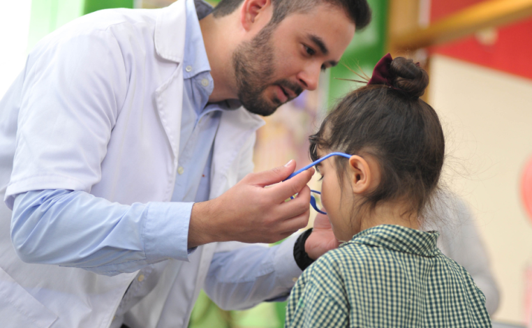Técnico óptico, con túnica, coloca lentes a una niña de unos cuatro años, con uniforme de centro de educación inicial