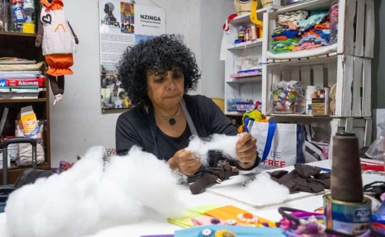 Mujer trabaja en un taller textil, manipulando algodón. Sobre la mesa en la que trabaja hay material textil. En los esta