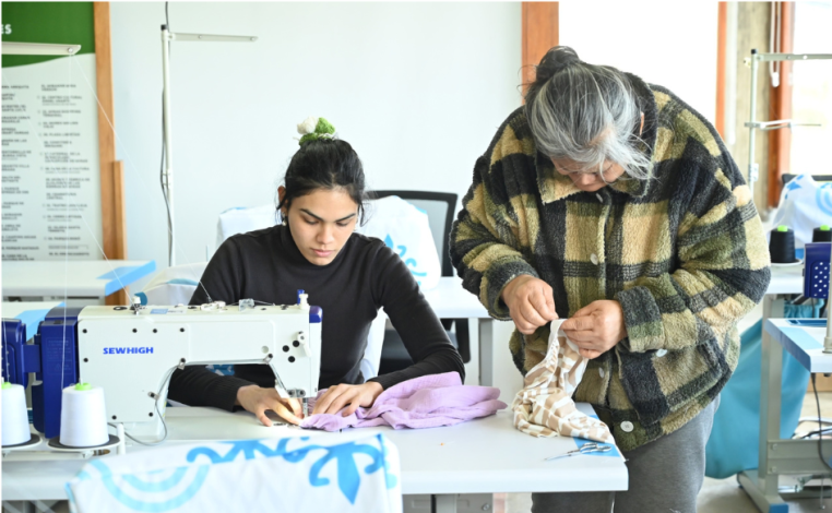 Mujer cosiendo sentada, con una máquina de coser, en el Centro de Costura Comunitario de la ciudad de Minas. A su lado, 