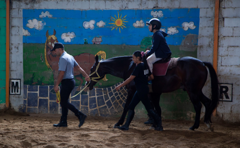 Niño con trastorno del espectro autista se traslada en caballo. A su lado, dos adultos lo acompañan de a pie. En el fond