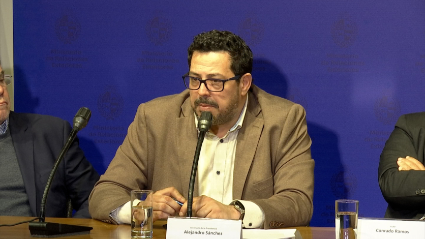 Secretario de Presidencia, Alejandro Sánchez, habla frente a un micrófono, sentado, con los antebrazos apoyados en una m