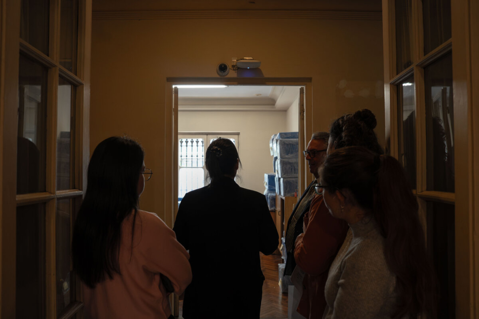 Referentes institucionales supervisan el traslado de archivos del pasado reciente. Cuatro personas conversan en un pasillo frente a una sala con cajas embaladas.