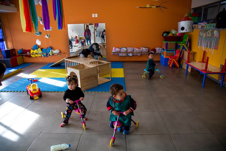 Dos niños y una niña juegan cada uno en su triciclo en sala de un año del CAIF