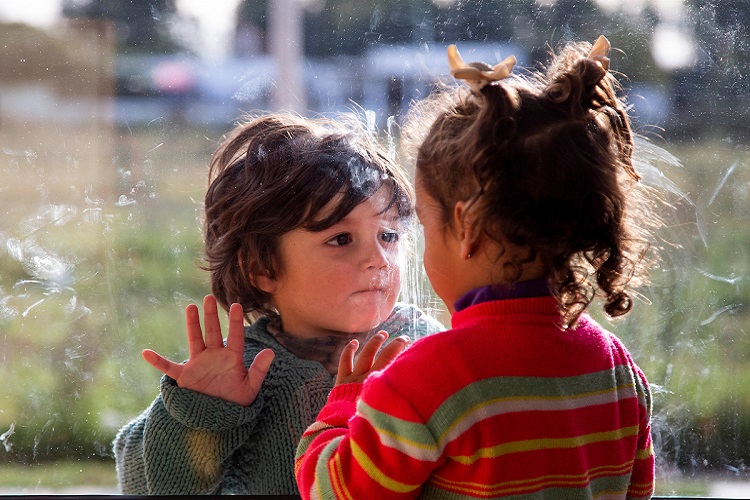 Un niño y una niña se miran y juntan sus manos a través de un vidrio