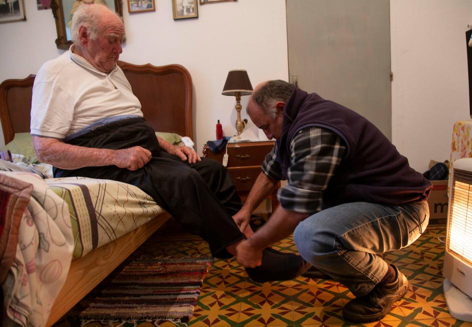 Cuidador ayuda en atar los cordones de su asistido. El cuidador está agachado y el asistido sentado en una cama.