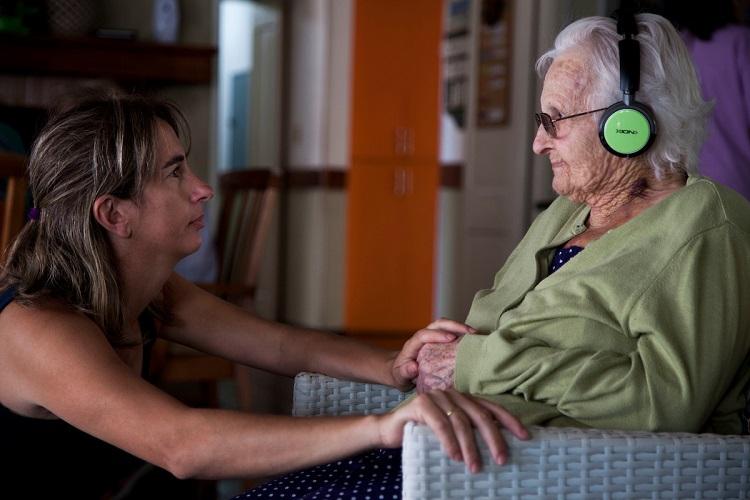 Cuidadora en centro de larga estadía mira a mujer mayor que está sentada con auriculares Cuidadora en centro de larga estadía mira a mujer mayor que está sentada con auriculares