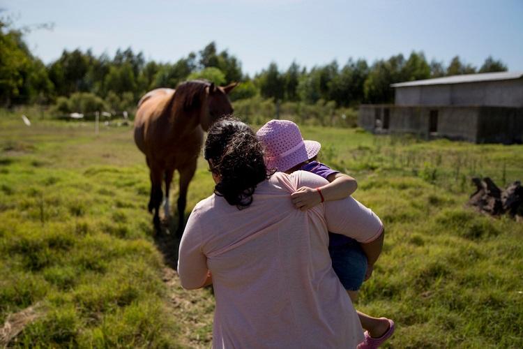 Cuidadora acompañando a una niña en su falda. Se ven de espaldas frente a un paiseaje de campo donde se ve un caballo.