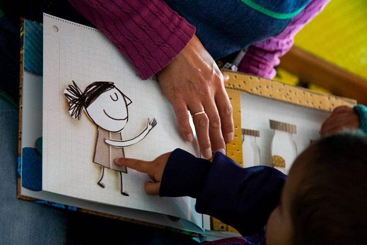 Se ven las manos de un niño pequeño en un CAIF y su educadora. Las manos de ambos están en el libro, el señala una niña dibujada. Se ven las manos de un niño pequeño en un CAIF y su educadora. Las manos de ambos están en el libro, el señala una niña dibujada.
