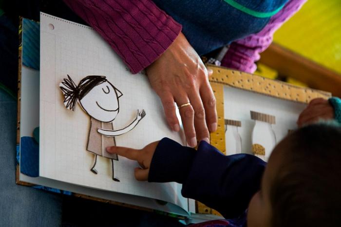 Se ven las manos de un niño pequeño en un CAIF y su educadora. Las manos de ambos están en el libro, el señala una niña dibujada.