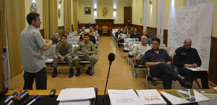 Personas en una jornada de planificación, militares, policías, civiles, hombres, mujeres, se ven libros en una mesa y un esquema en un papelografo en la pared.