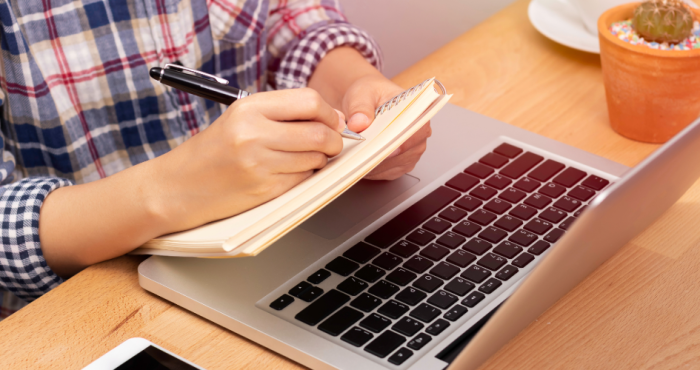 Imagen ilustrativa: primer plano de una mano escribiendo un papel al lado de una laptop