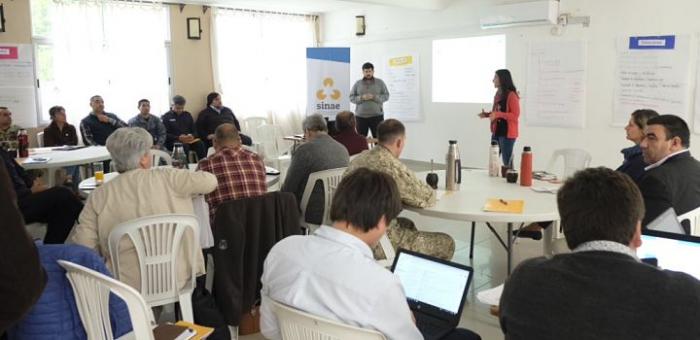 Imagen de taller de GIR, con personas sentadas y una mujer y un hombre hablando al frente con papelógrafos detrás.