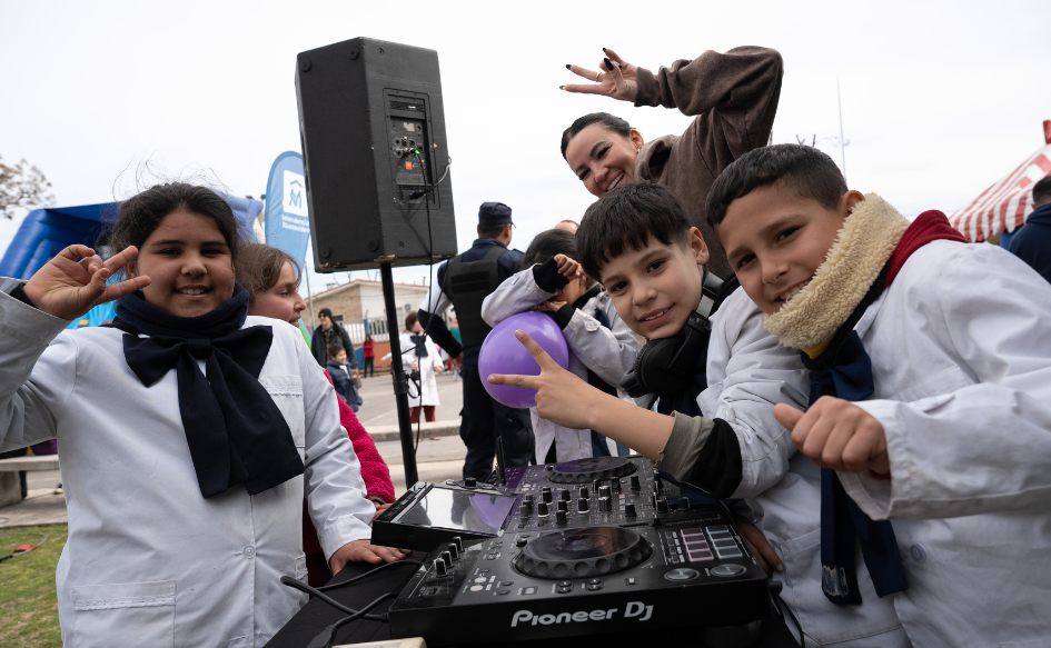 Dos niños y una niña saludan a la cámara junto a una consola de sonido durante la celebración del Día de la Niñez en Cerro Norte