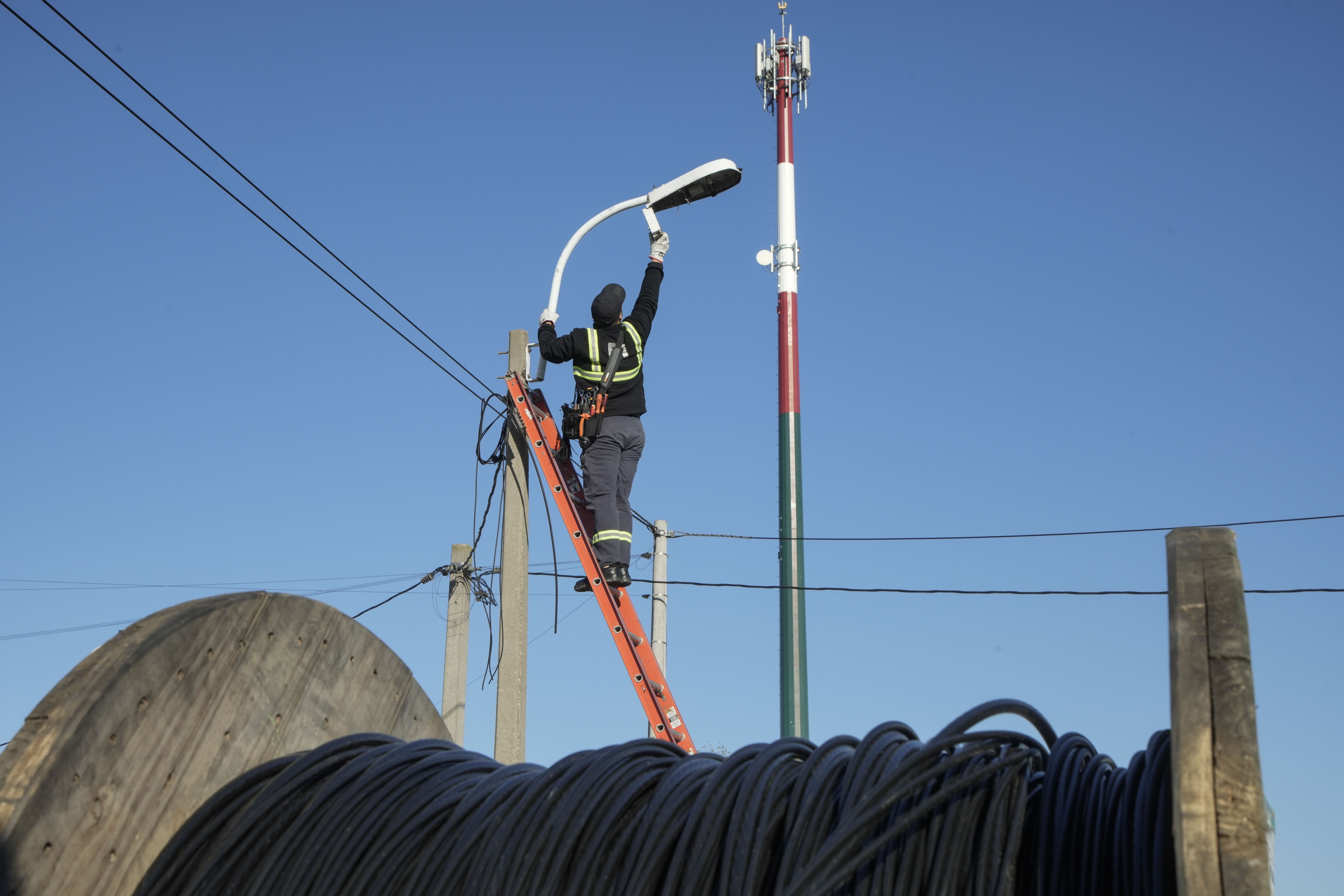 Un operario trabaja en la instalación de una luminaria de alumbrado público sobre una escalera, con una gran bobina de cable en primer plano y una antena de fondo.