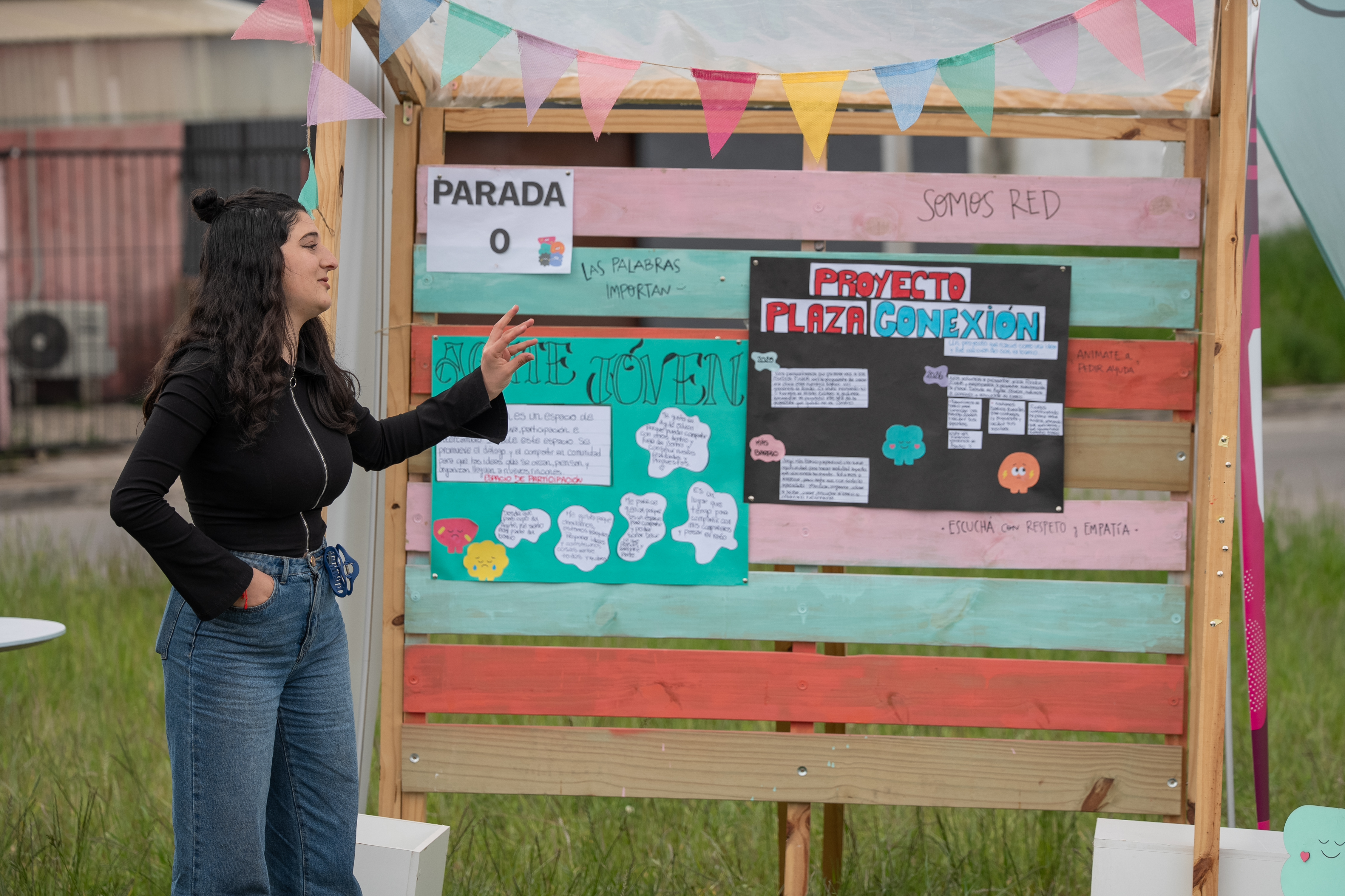 Participante de Ni Silencio Ni Tabú habla señalando cartelería durante el taller participativo de diseño de la plaza conexión. En la cartelera, se observan cartulinas con la leyenda "Agite Joven" y "Proyecto Plaza Conexión"