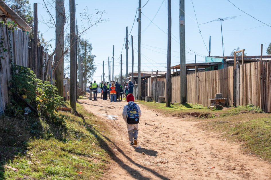 Niño con mochila camina por una calle de tierra; al fondo, personas con chalecos.