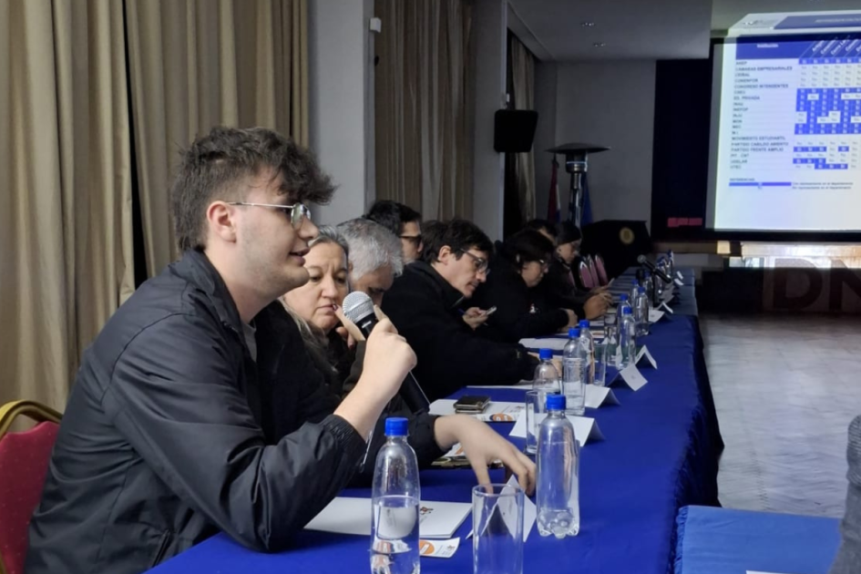 Personas sentadas en reunión y una persona hablando mientras sostiene micrófono.