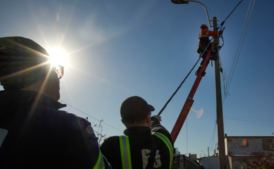 Trabajador repara el tendido eléctrico desde una escalera, asistido por dos compañeros.