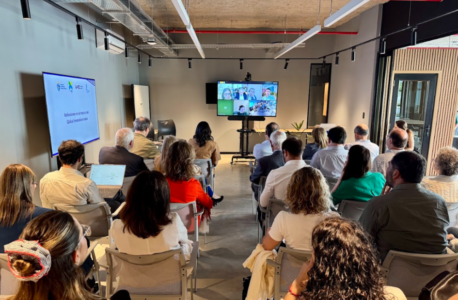 Público presente sentado en sillas. Delante, una tv con los expositores den una videollamada